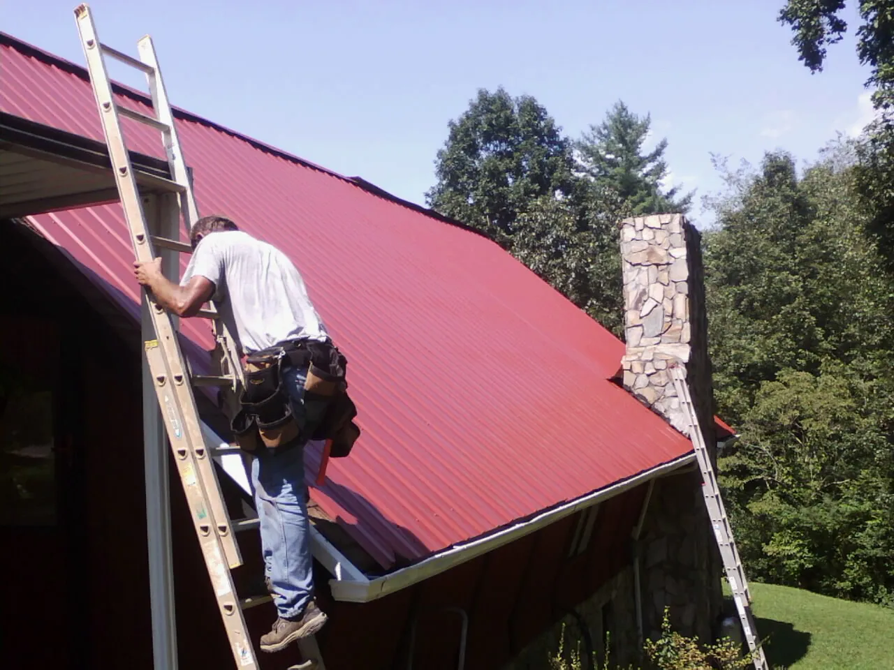 Roofer on a ladder beside a red metal roof and stone chimney — Soffit & Fascia Repair by Roofing Brothers in Christiansburg
